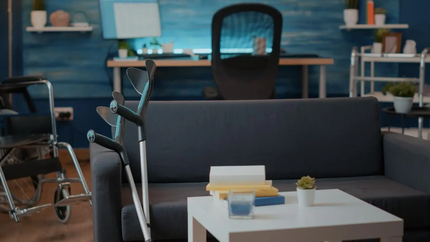 Medical crutches on a couch, used by a person with a physical disability. An empty living room with walking support sticks and a wheelchair to provide aid and accessibility for transportation.