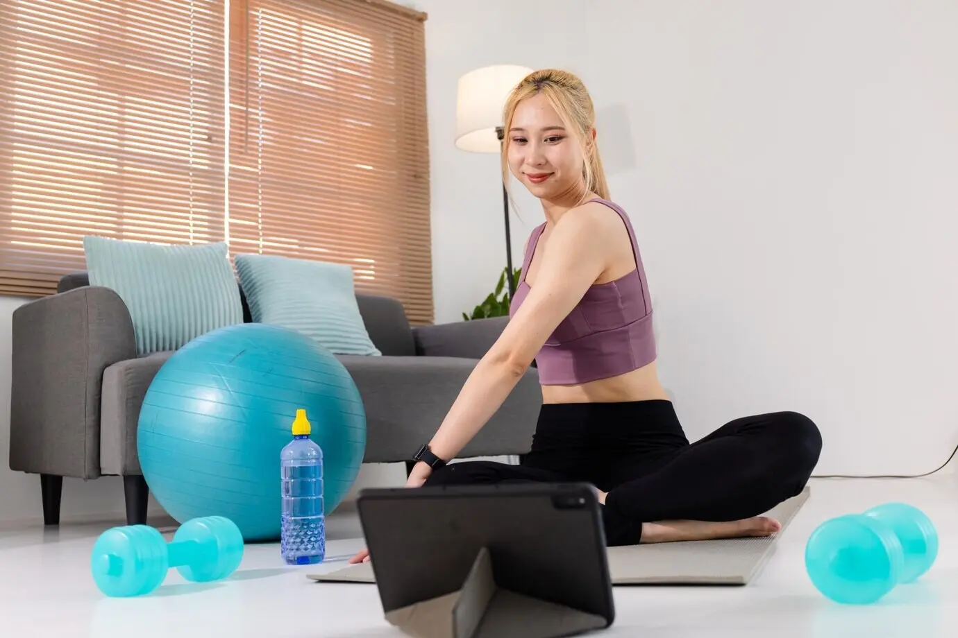 A pretty young Asian woman in sportswear sits on an exercise mat, watching online training videos on a digital tablet.